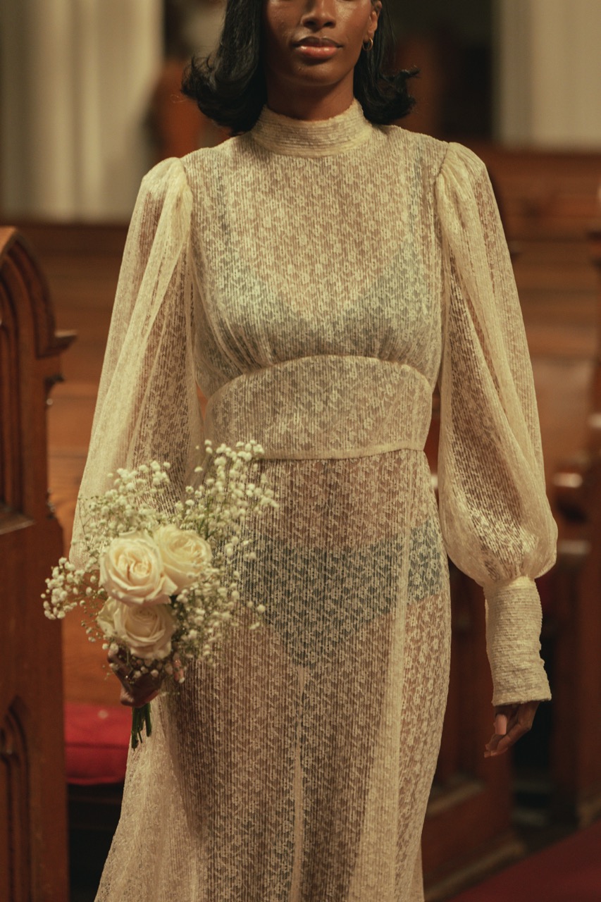 Model walks down the aisle carrying a bouquet of white roses during Tanner Fletcher's bridal fashion show at New York Bridal Fashion Week, wearing a sheer long-sleeve bridal gown inside a historic church.